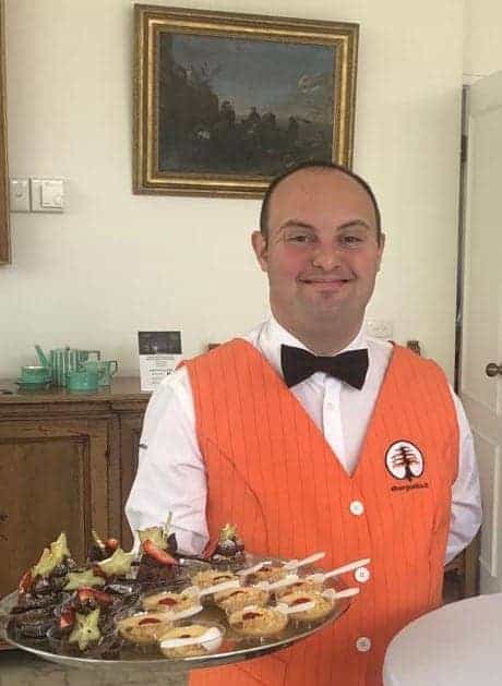 A person in an orange vest and bow tie smiles, holding a tray of appetizers in a room with a painting on the wall.